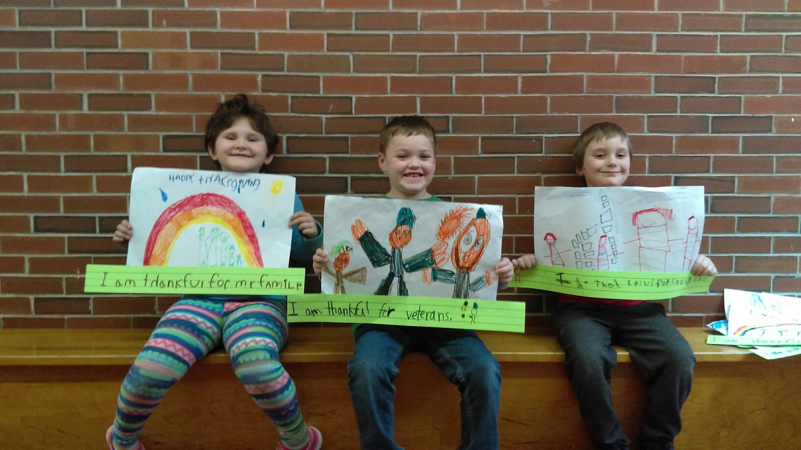 3 1st graders holding illustrations they drew captioned with "I am thankful for ..." sentences